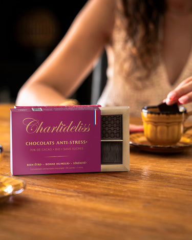 Femme brune trempant un carré de chocolat Charlidéliss dans sa tasse de café en face d'une autre personne qui fait de même. Le coffret de chocolat est posé au milieu et on voit très clairement indiqué ses bienfaits anti-stress naturels puissants.