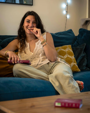 Femme brune aux cheveux bouclés, souriante, assise sur un canapé bleu mangeant des chocolats Charlidéliss pour bénéficier de leur effet anti-stress naturel puissant.